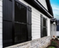 Close-up view of the side of a modern residential house featuring a combination of vertical white siding and stone veneer at the base. The house has large windows with dark frames and shutters, enhancing the stylish contrast against the white siding